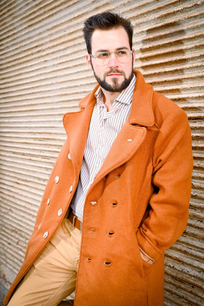Homme portant des lunettes Loumy, manteau orange et chemise rayée, posant contre un mur en métal ondulé