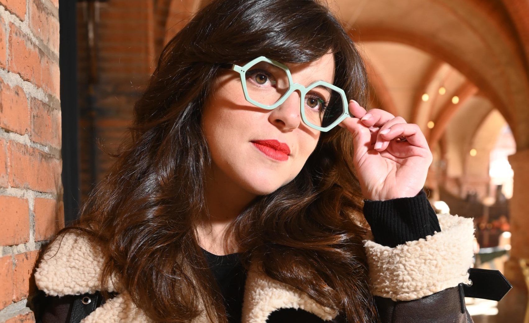 Portrait rapproché d’une femme portant des lunettes Loumy vert clair, devant des arcades en briques